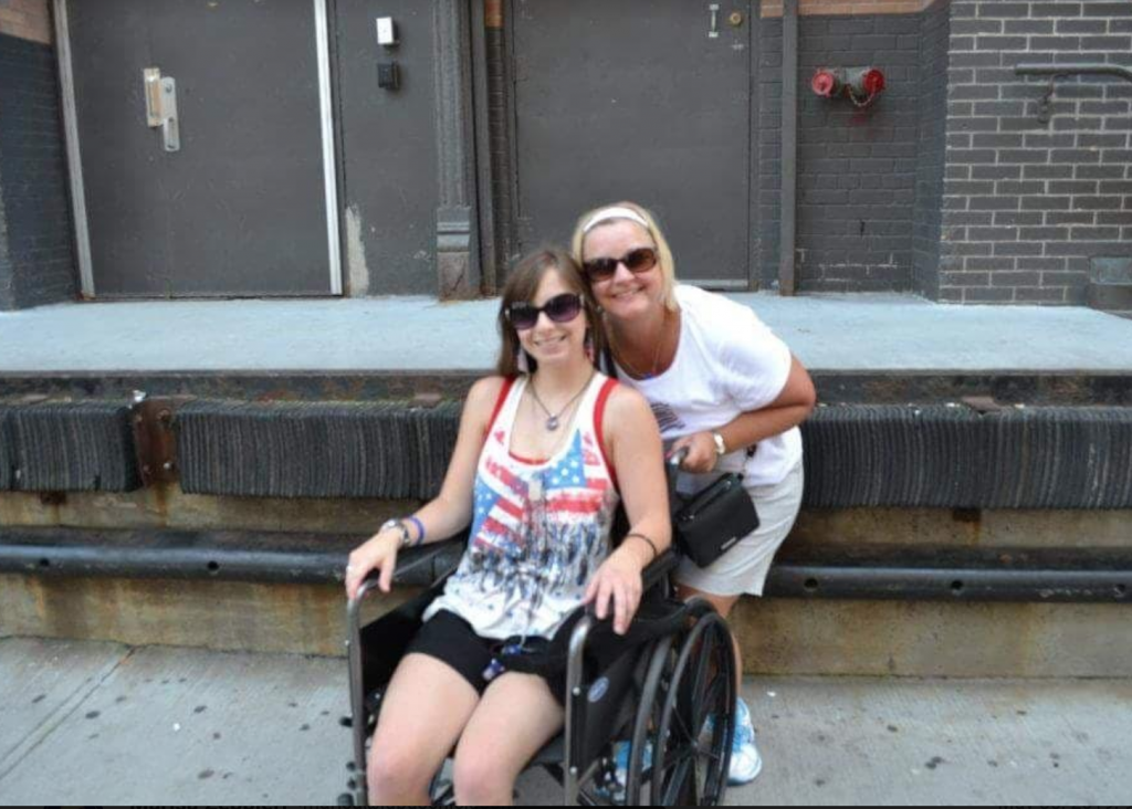 A photo of a young woman in a wheelchair with her friend behind her. 