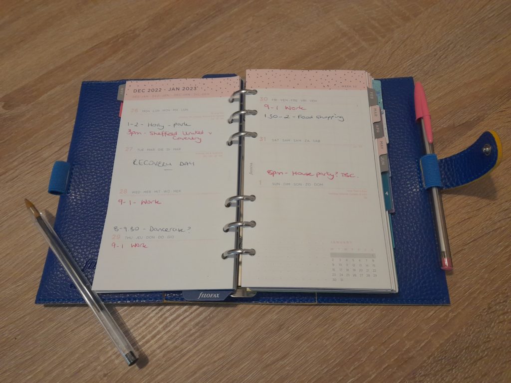 A simplified version of Katey's Filofax that helps her fatigue. It is a blue planner with several tabs. The pages are white with pink speckled headings. 
